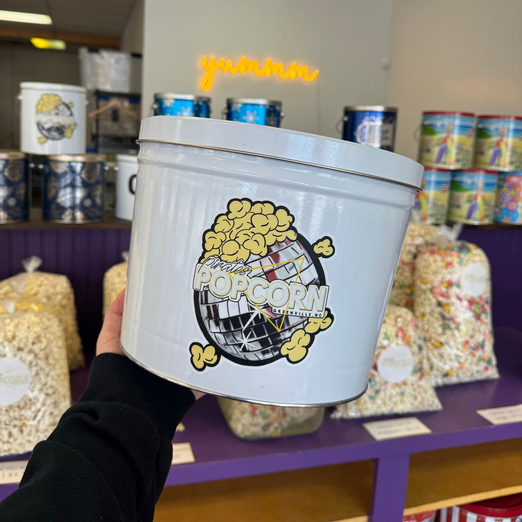 A person holding a white 2-gallon popcorn tin with a graphic design of popcorn and a pirate ship on the front.