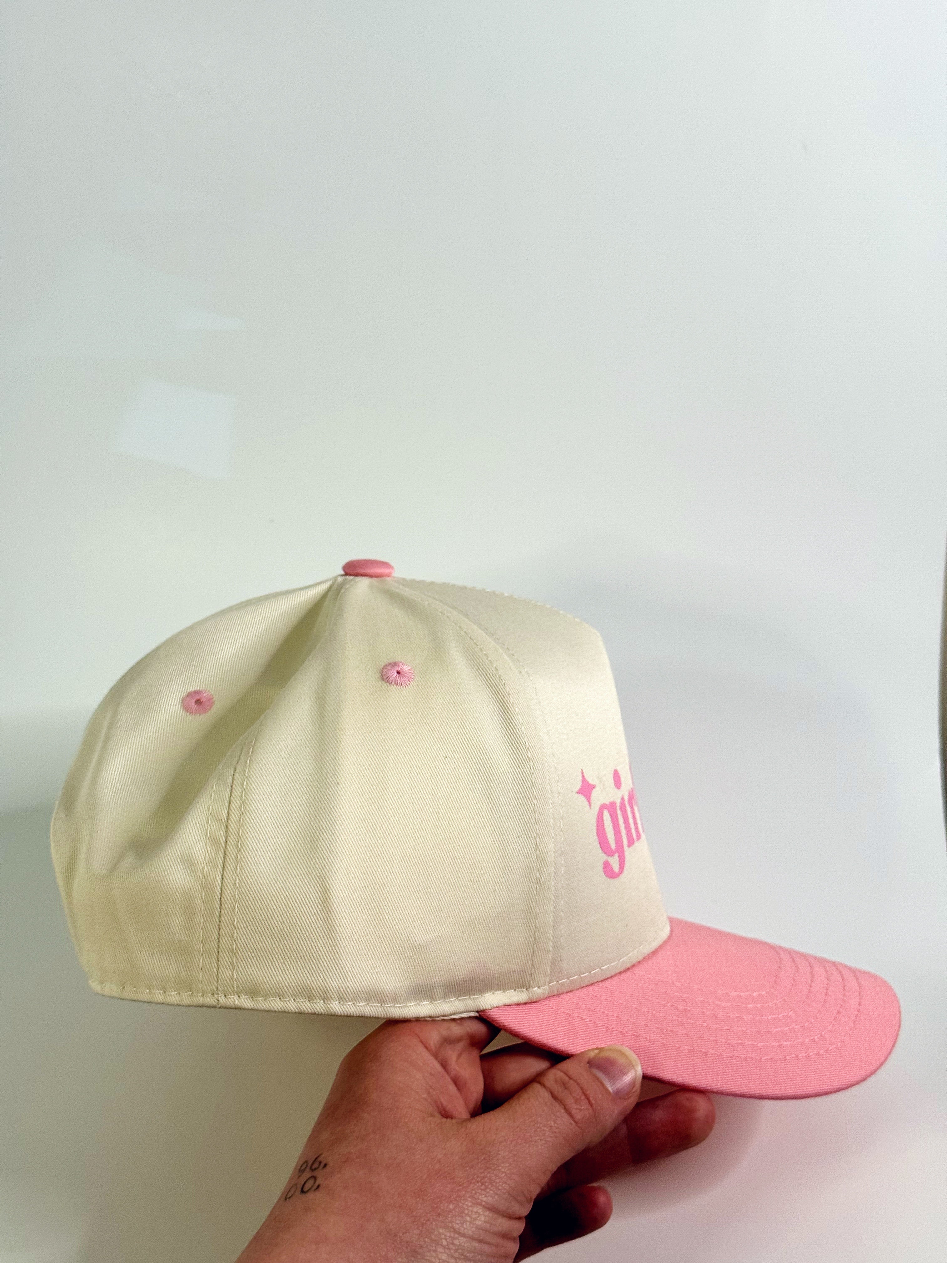 Two baseball caps, one beige with pink visor and one pink with pink visor, held by a hand against a light background.