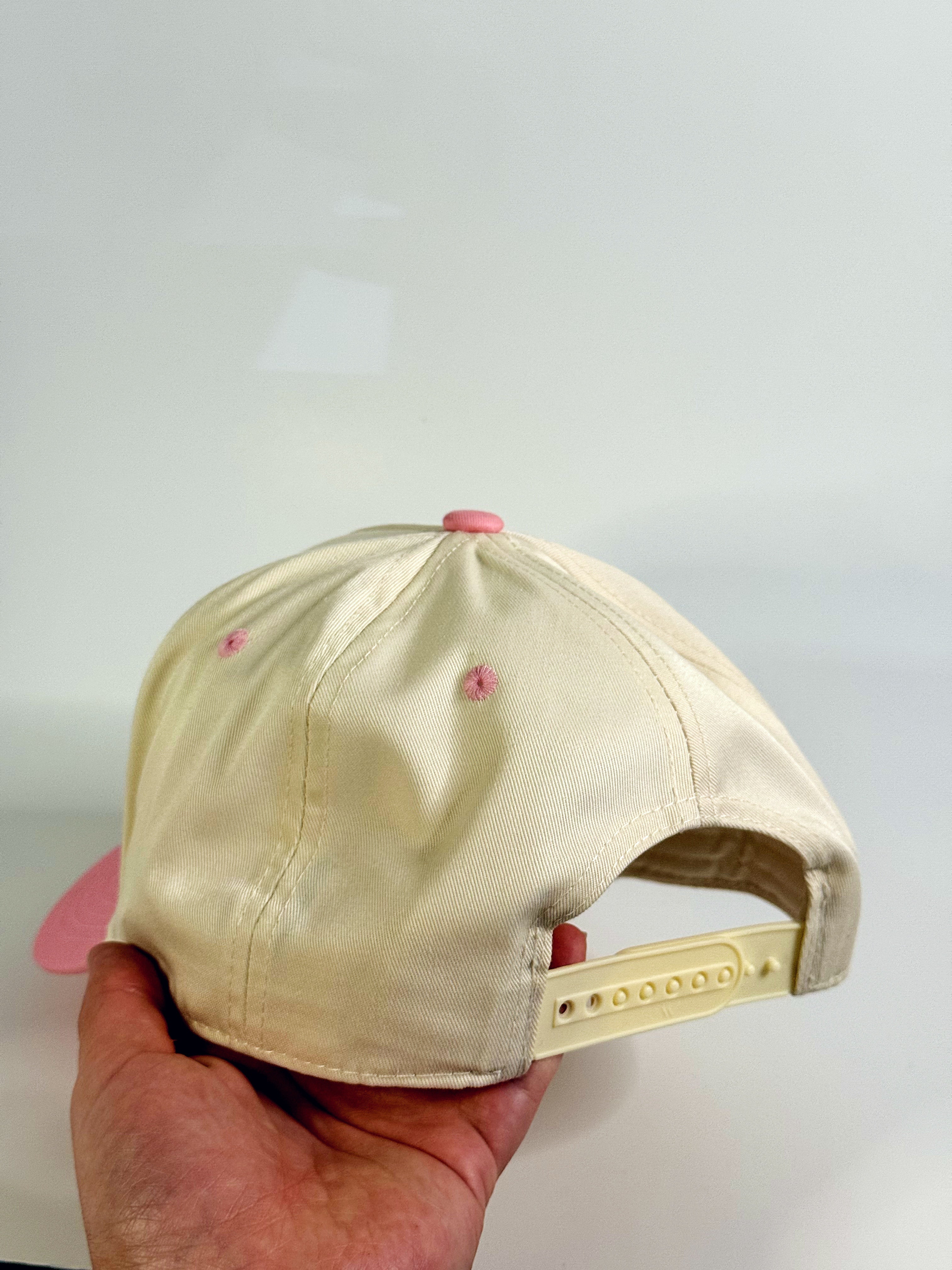 Beige cap with pink visor held by a hand against a light gray background