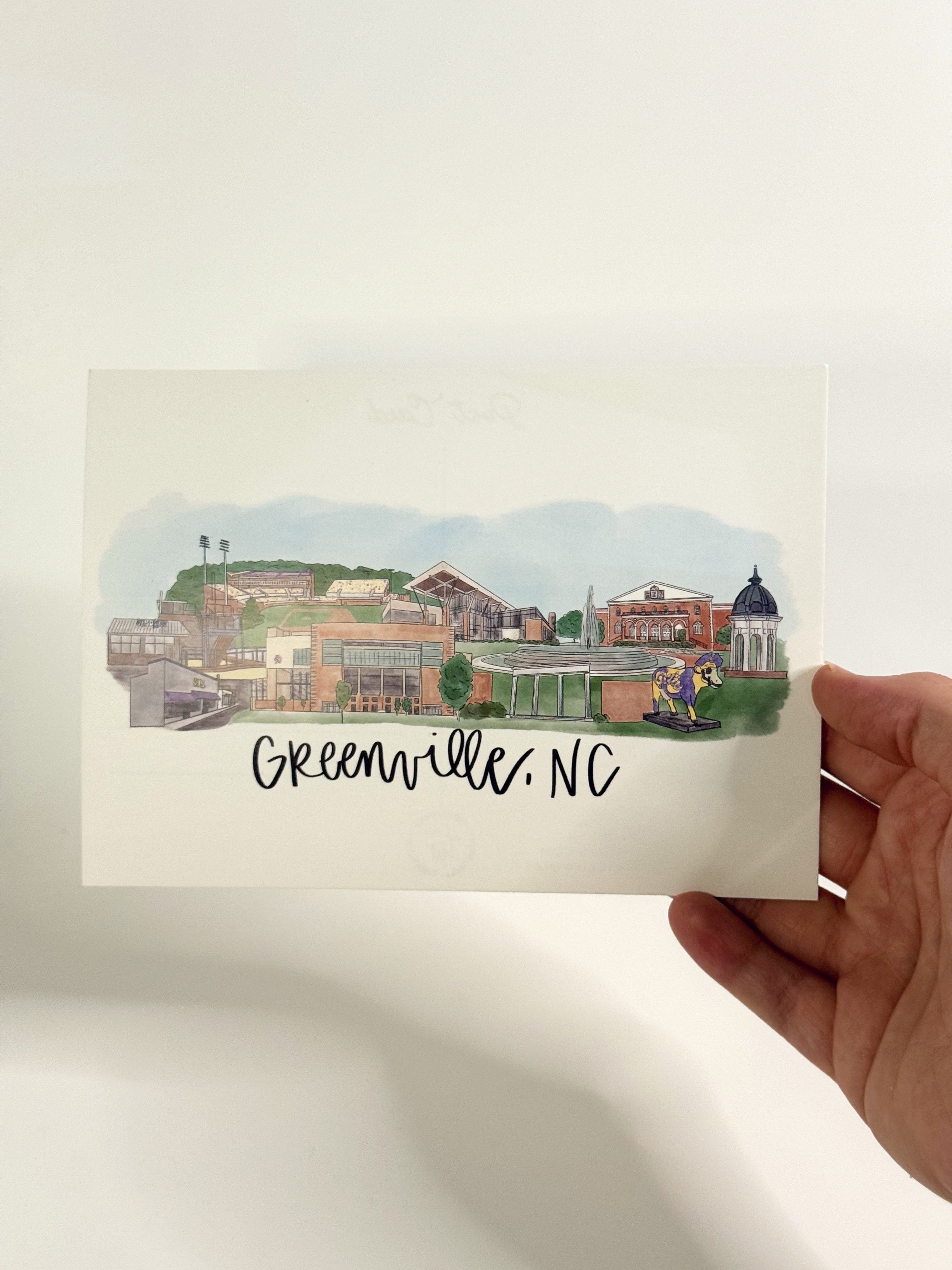 Hand holding a card with a watercolor illustration of Greenville, NC on a plain background